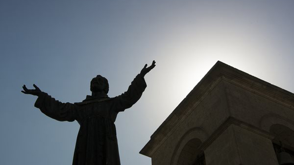 A statue of St. Francis with outstretched arms and the sun behind him
