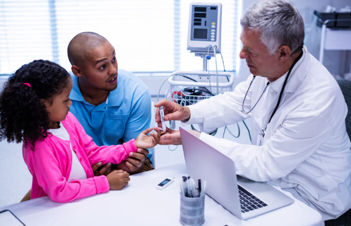 Doctor checking young girl's blood sugar. Dad is sitting with her asking the doctor questions. 