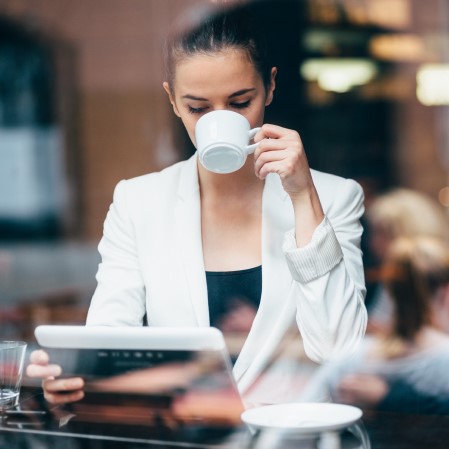 A women in white drinks coffee in a coffee shop while reading a tablet