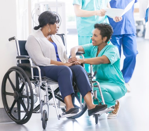 Photo of female nurse kneeling and holding the hand of a female patient in a wheelchair