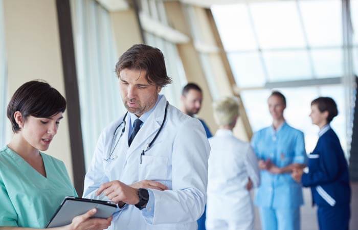 2 doctors in the foreground looking at patient chart while a group of doctors in the back have conversation