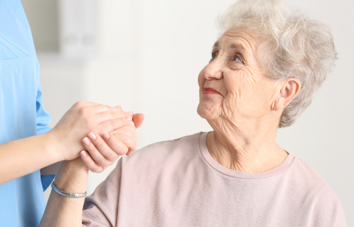 An older women sits while holding the hand of a standing clinician wearing blue