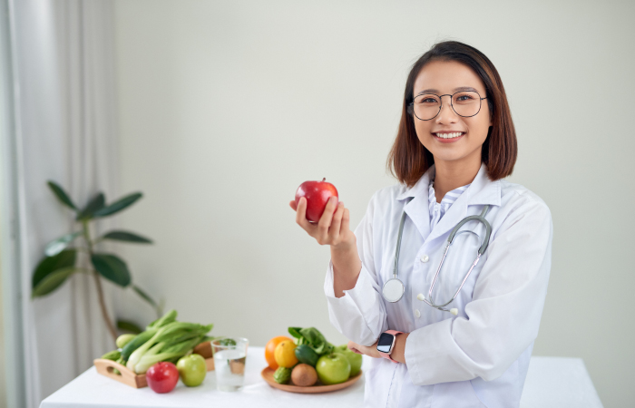 female asian doctor standing in front of a table full of fruit holding an apple and smiling at the camera 