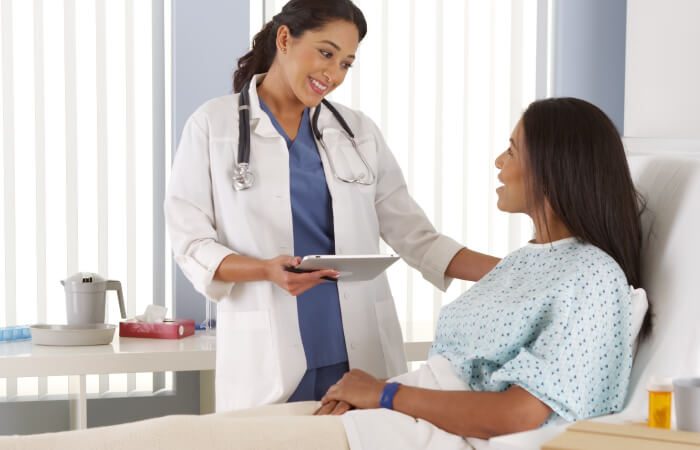 A female doctor stands at the bedside of a female patient touching her shoulder and holding a tablet