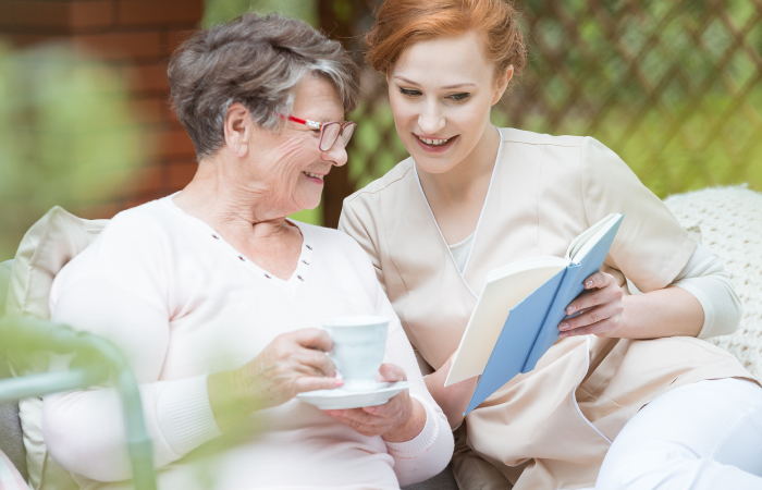 Older women drinks coffee on a park bench as a younger female care provider reads a book