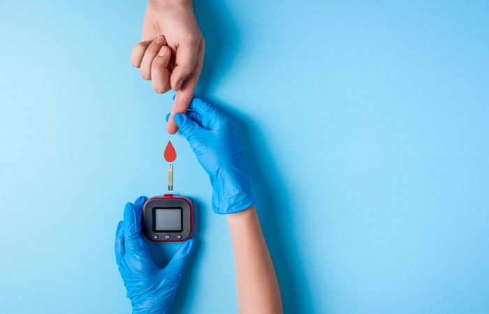 A technician wearing blue gloves assists a patient prick their finger to check blood sugar levels