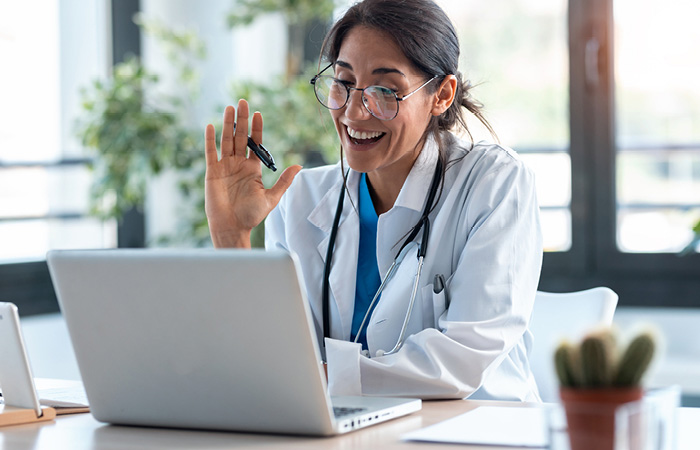 Young female doctor with glasses waving to patient on laptop screen