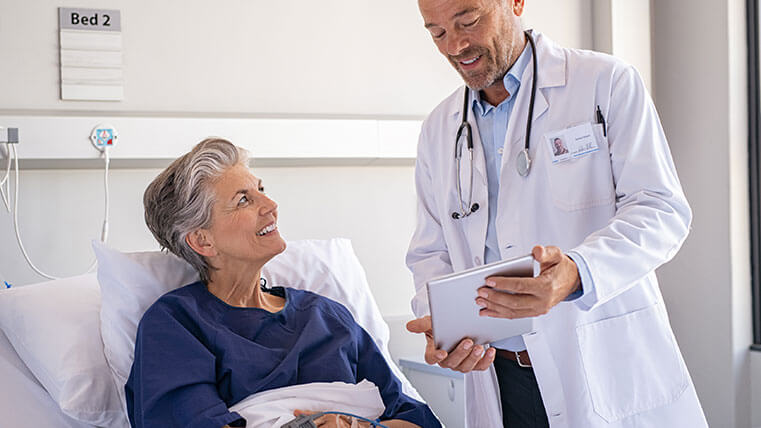 Mature female patient in a blue gown sitting in hospital bed looking up smiling to her male doctor standing bedside.