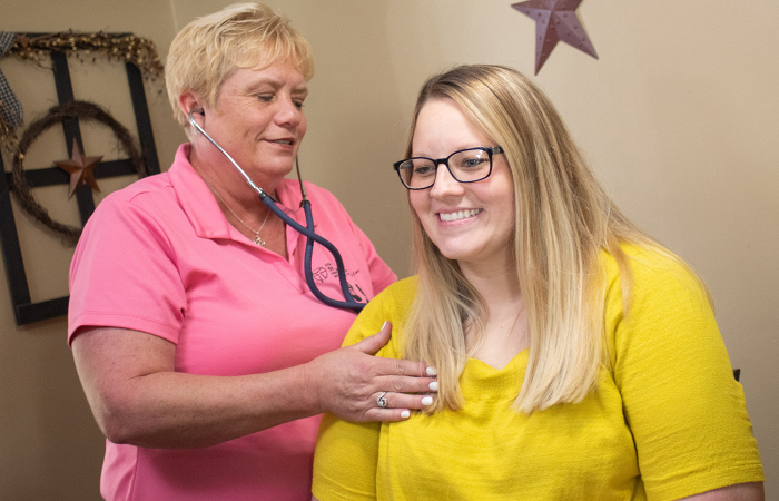 female nurse in pink shirt checking the heartbeat of woman in yellow shirt with glasses