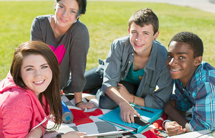 Group of teens, two girls and two boys sitting on picnic blanket and smiling at the camera