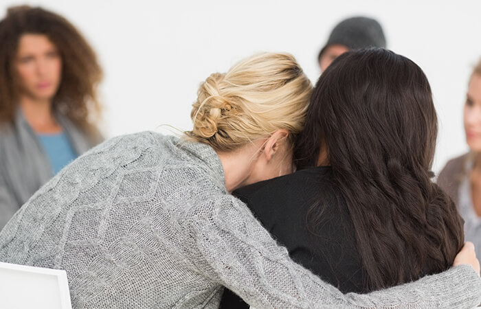 Diverse group of women at a support group sitting in a circle. Closest to the camera is a woman leaning in to hug another.