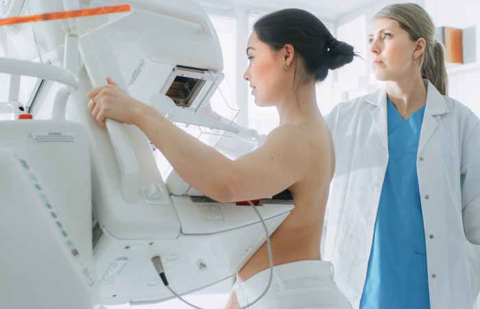 Young woman standing in front of mammogram machine and female doctor standing behind her