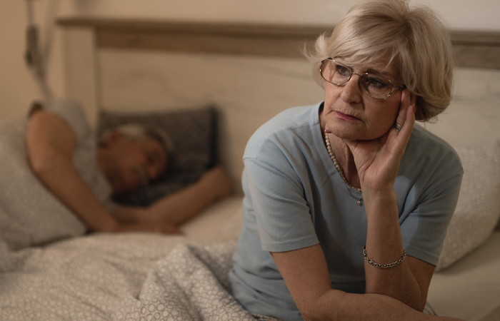 Elderly woman sitting on edge of the bed while elderly man sleeps in bed