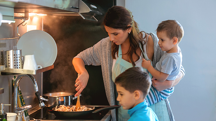 Mother holding toddler and cooking with older son looking on