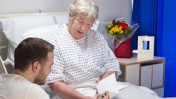 Senior woman in hospital with young man reading her a greeting card
