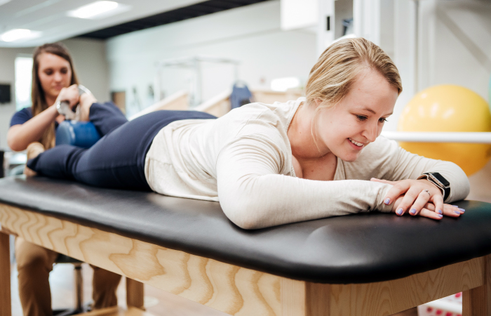 A female patient lays on her stomach on a trainers table as a trainer works on her ankle. 