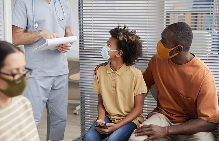 A black father and his teenage son both with masks on speaking to a male nurse