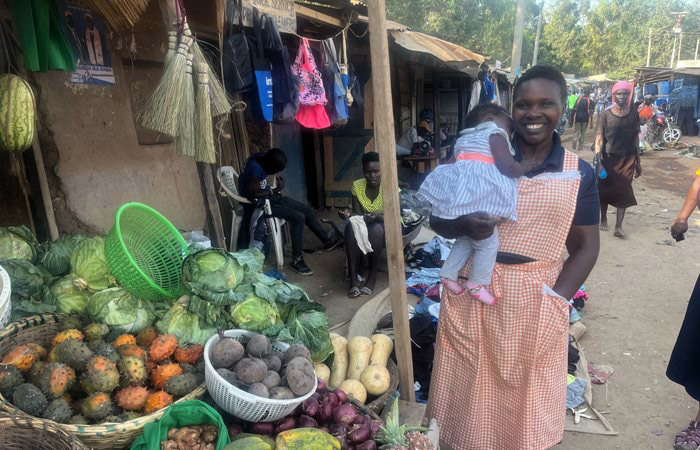 An African women holds an infant and stands in a market behind fresh produce.