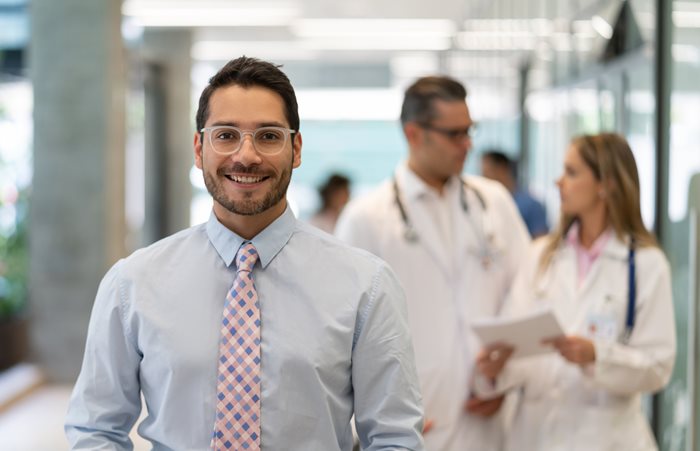 Professional man wearing dress shirt and tie smiles as he walks down hallways in front of medical providers