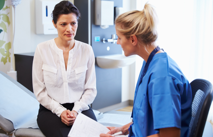 blonde female nurse speaking to a brunette female patient sitting on edge of hospital bed 