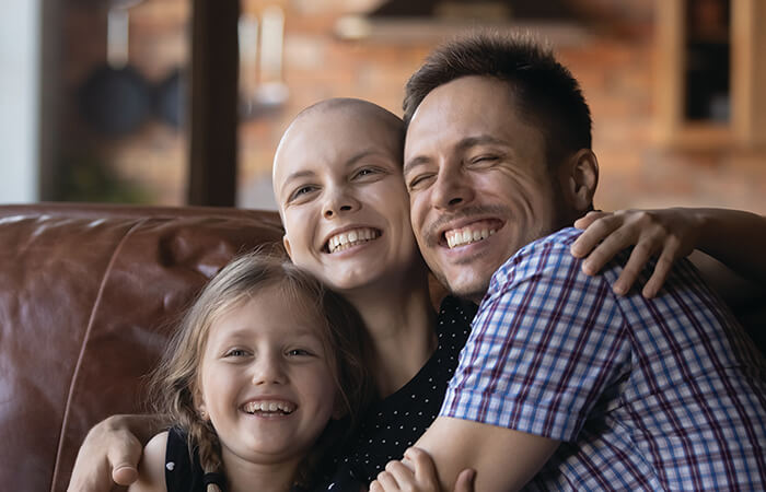 A young women with a shaved head smiles and hugs her family on their sofa