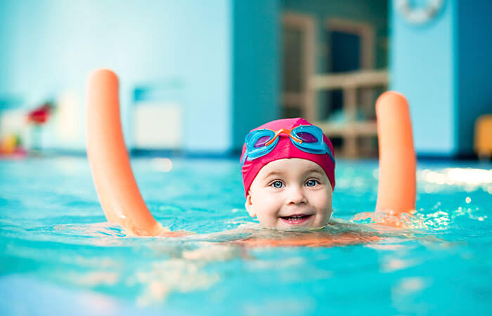 young child in pink swimming cap in the swimming pool with orange pool noodle