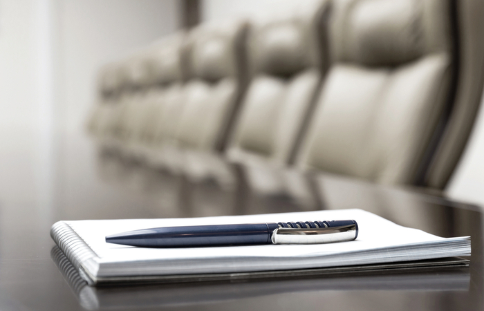 A pen and notepad sit on top of a board room table surrounded by chairs
