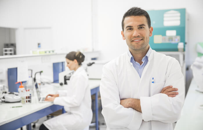 Man in lab coat smiles while female in lab coat works on a computer in the background